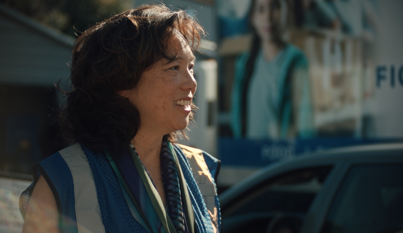 Kathy Kwan wears a reflective safety vest and a scarf smiles while standing outdoors. 