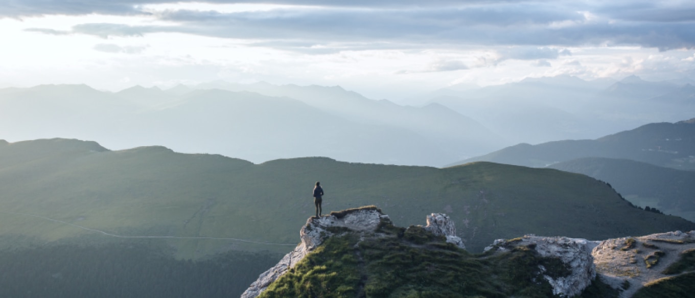 A lone hiker stands on the edge of a rocky peak, overlooking a vast expanse of rolling green hills and distant mountain ranges. The sky is filled with soft clouds, and the sunlight casts a hazy glow over the landscape