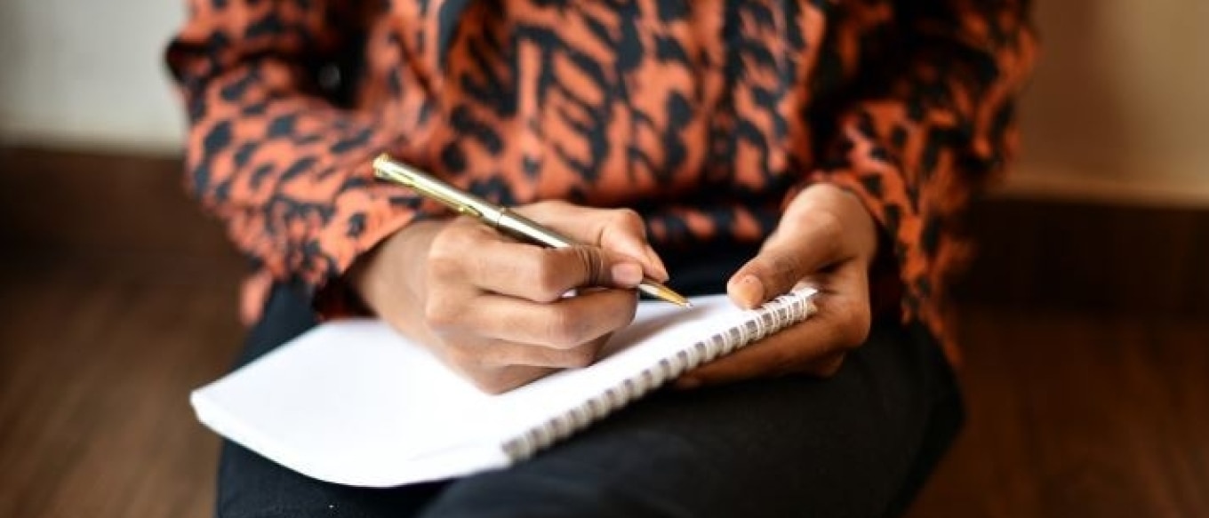Close-up of a person wearing an orange and black patterned shirt, holding a gold pen and writing in a spiral-bound notebook while sitting on a wooden floor.