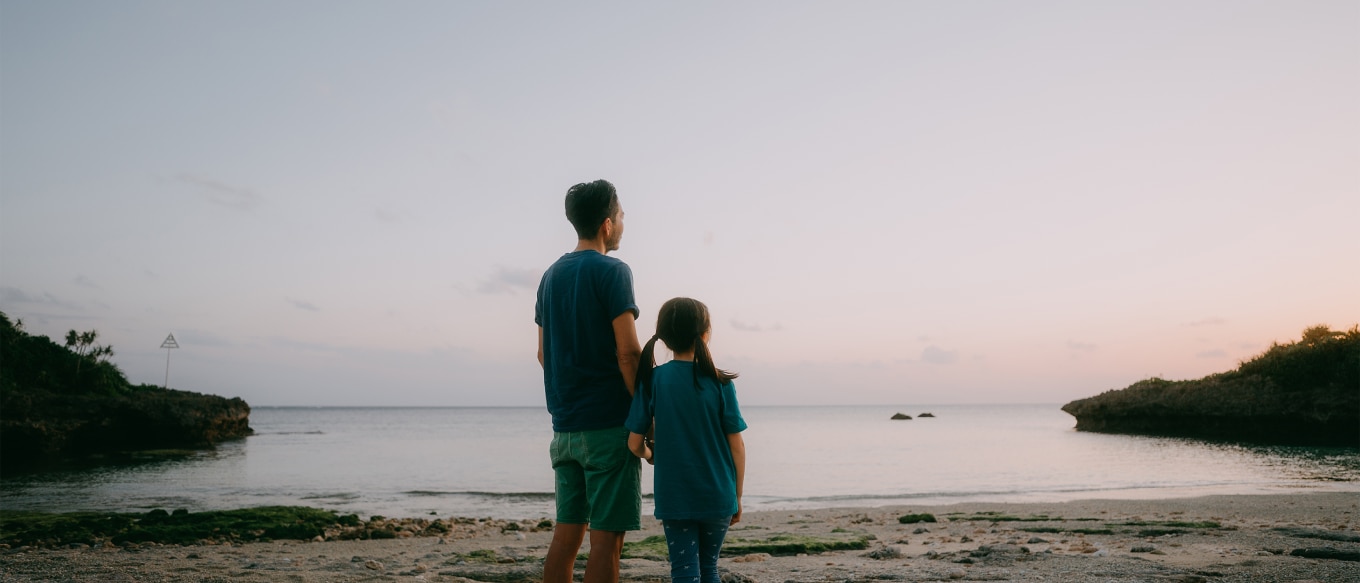 A father and his young daughter stand on a sandy beach at sunset, gazing out at the calm ocean. They are both wearing matching blue shirts, and the girl holds her father's hand. The sky displays soft pastel hues, and rocky outcroppings frame the shoreline
