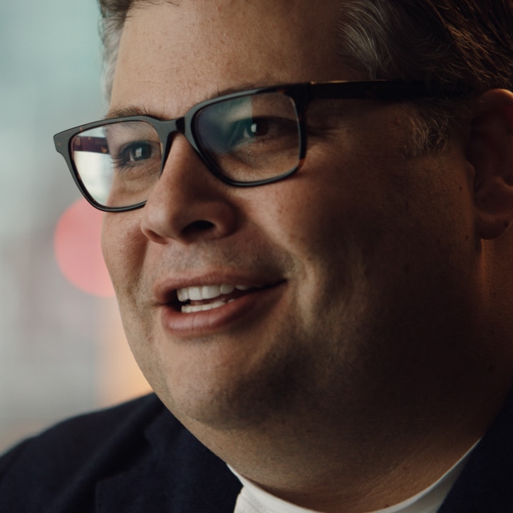 A close-up portrait of a RJ Melman wearing black-framed glasses and a dark blazer. He has a friendly expression with a slight smile, and his eyes are focused on something off-camera. The blurred background features warm, ambient lighting.