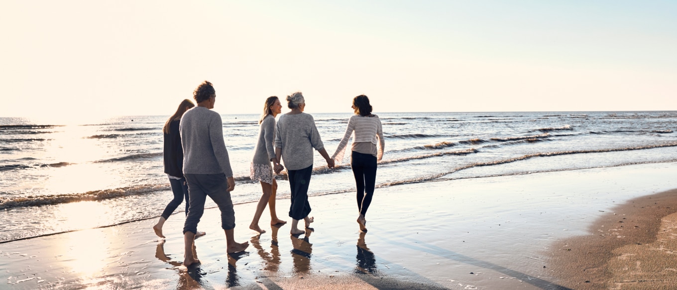 A group of five people, including multiple generations, walk barefoot along the shoreline at sunset. They are dressed in light sweaters and casual clothing, holding hands and enjoying the peaceful ocean waves. The setting sun casts long shadows on the wet sand, creating a warm and reflective atmosphere