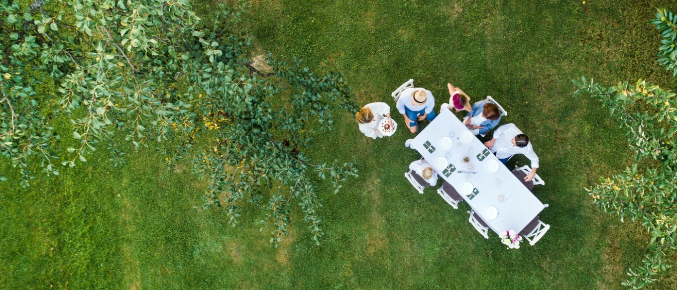 Aerial view of a small group of people gathered around a long, elegantly set dining table on a lush green lawn. The table is covered with a white tablecloth and decorated with plates, napkins, and floral arrangements. The surrounding trees create a natural canopy, and one person is approaching the table carrying a cake