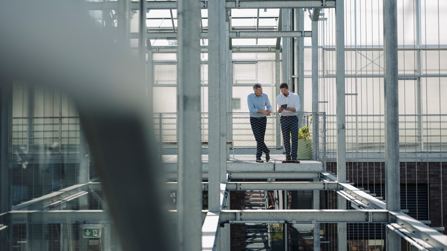 One man shows another man his notebook as they stand by a fence in front of the steel frame of a building under construction. 