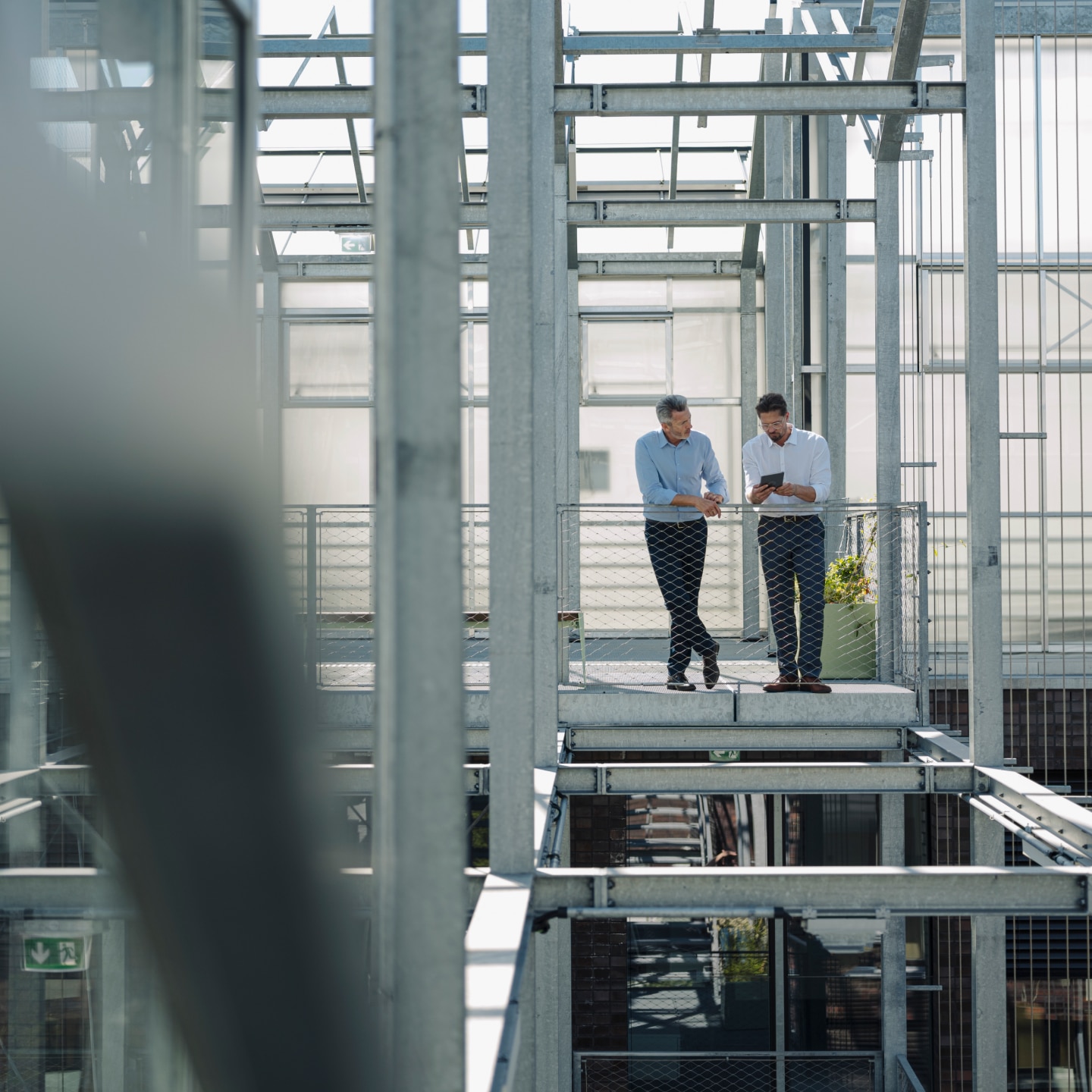 One man shows another man his notebook as they stand by a fence in front of the steel frame of a building under construction.
