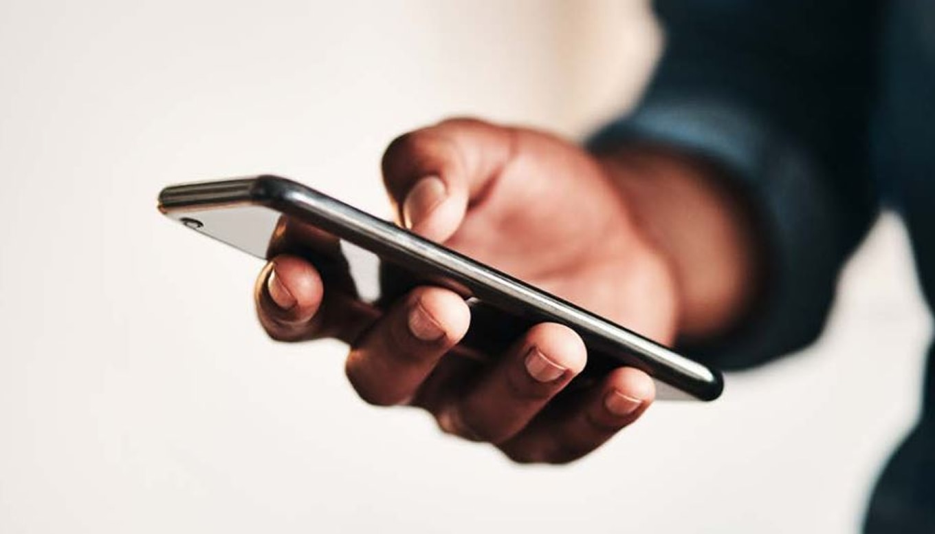 A close-up of a person's hand holding a modern smartphone. The phone has a sleek design with a reflective surface, and the person is wearing a dark-colored long-sleeve shirt. The background is blurred