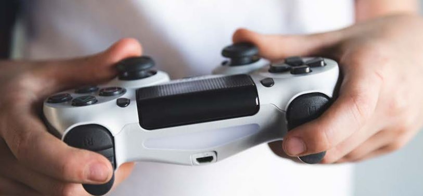 A close-up of a person holding a white and black gaming controller with both hands. The fingers are positioned on the buttons and thumbsticks, indicating gameplay. The background is blurred, and the person is wearing a white shirt