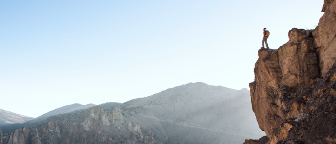 A hiker wearing a backpack stands on the edge of a rocky cliff, gazing into the distance. The background features rugged mountains and a clear sky.