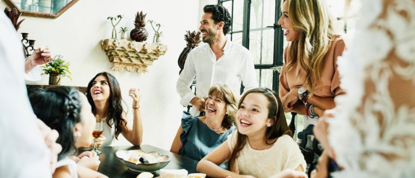 A joyful family gathering in a warmly lit dining area. Several people of different ages are laughing and smiling while engaging in conversation. A young girl in the foreground is grinning, while others around the table share a lively moment. The room is decorated with rustic and tropical elements, with natural light streaming in from large windows