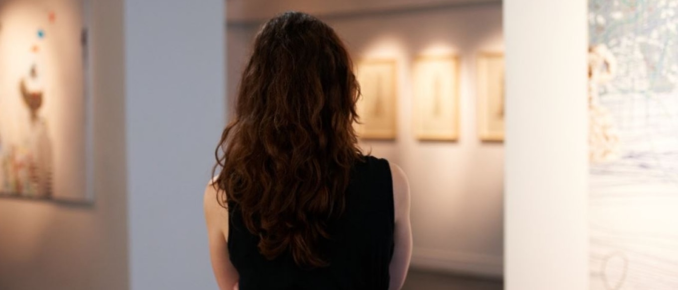 A woman with long, wavy brown hair wearing a black sleeveless top stands in an art gallery, viewed from behind. She is looking at framed artworks on a softly lit wall, surrounded by modern white pillars and additional paintings in the background.