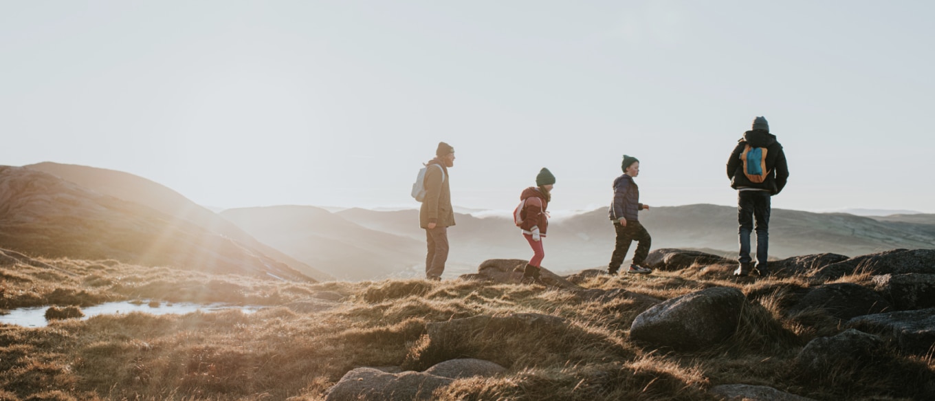 A group of four hikers walking on a grassy, rocky landscape during sunrise or sunset. The group includes adults and children, all wearing warm clothing and backpacks, enjoying the scenic view of rolling hills under a clear sky.
