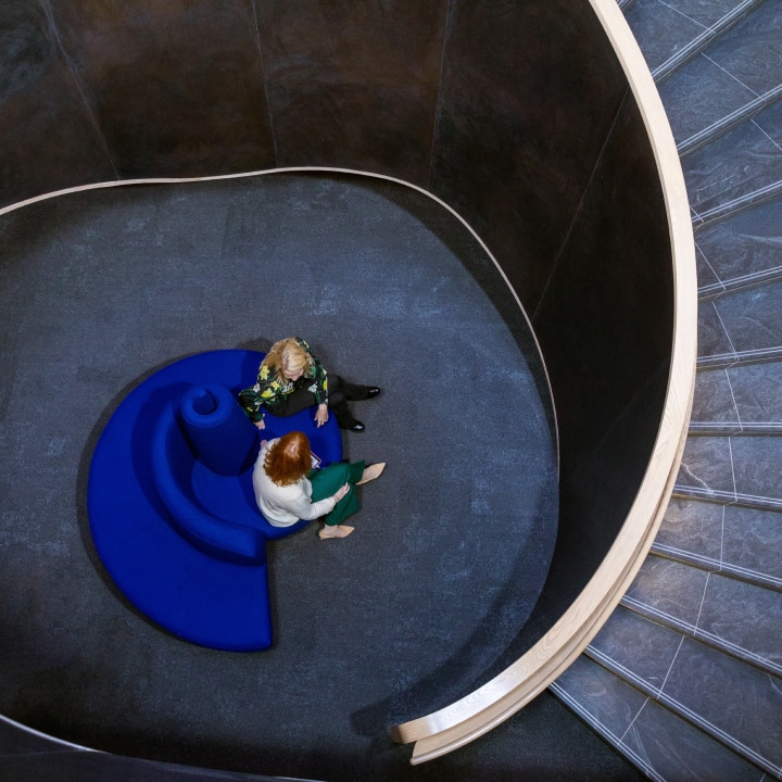 A high-angle shot captures a spiral staircase with two women seated on a blue, snail-shaped couch. The staircase features dark gray walls and gray steps with a white handrail. Small lights line the wall along the spiral. The women, one with blonde hair and one with red hair, are facing each other.