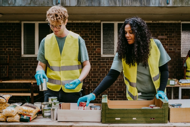 Two volunteers wearing reflective vests and gloves sorting canned goods at an outdoor food distribution table.