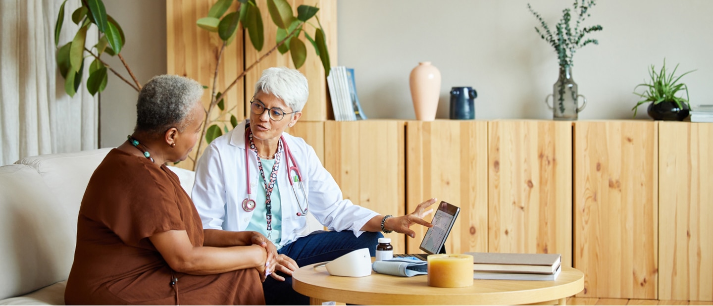 A doctor with a stethoscope shows a tablet to an elderly woman on a couch. Medical tools and books sit on a table in a cosy, well-lit room.