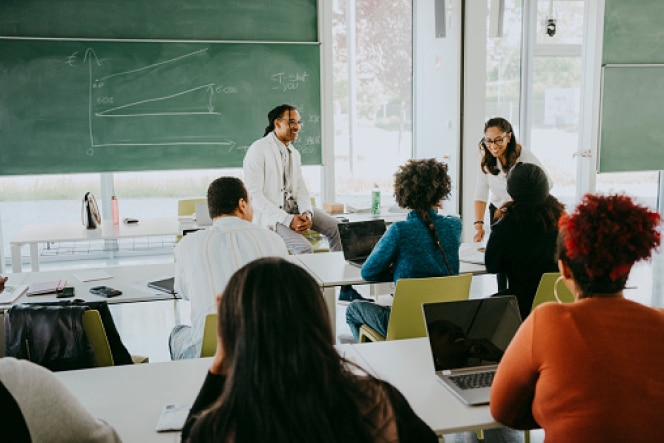 A diverse group of students in a classroom listening to a discussion led by two instructors near a chalkboard.