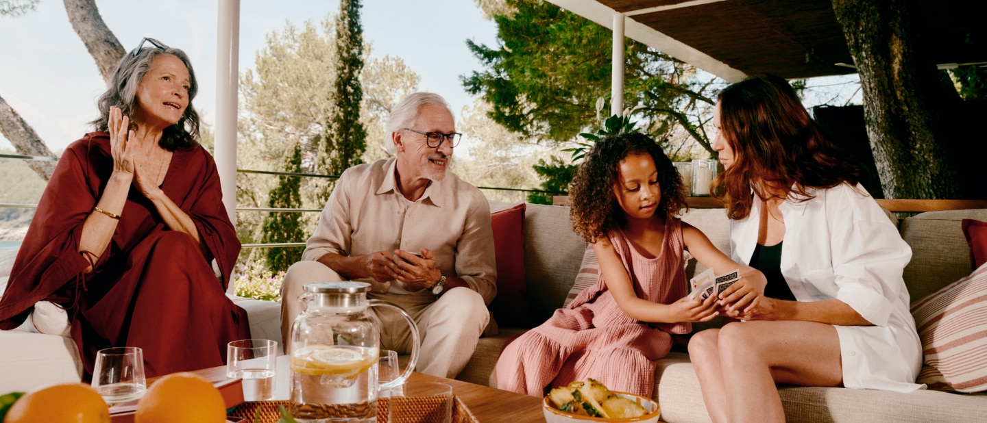 A multigenerational family relaxes on a patio. An elderly couple smiles while a young girl and a woman play cards. A table holds drinks and fruit, with lush greenery in the background