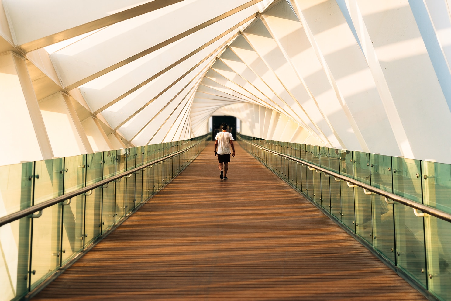A person walks along a modern, enclosed pedestrian bridge with geometric white beams and glass railings. Sunlight casts angular shadows on the walls, creating a dynamic pattern