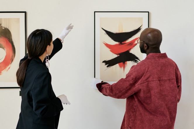 Two people view and discuss abstract art in a gallery, with one pointing toward a framed painting featuring bold red and black brushstrokes.