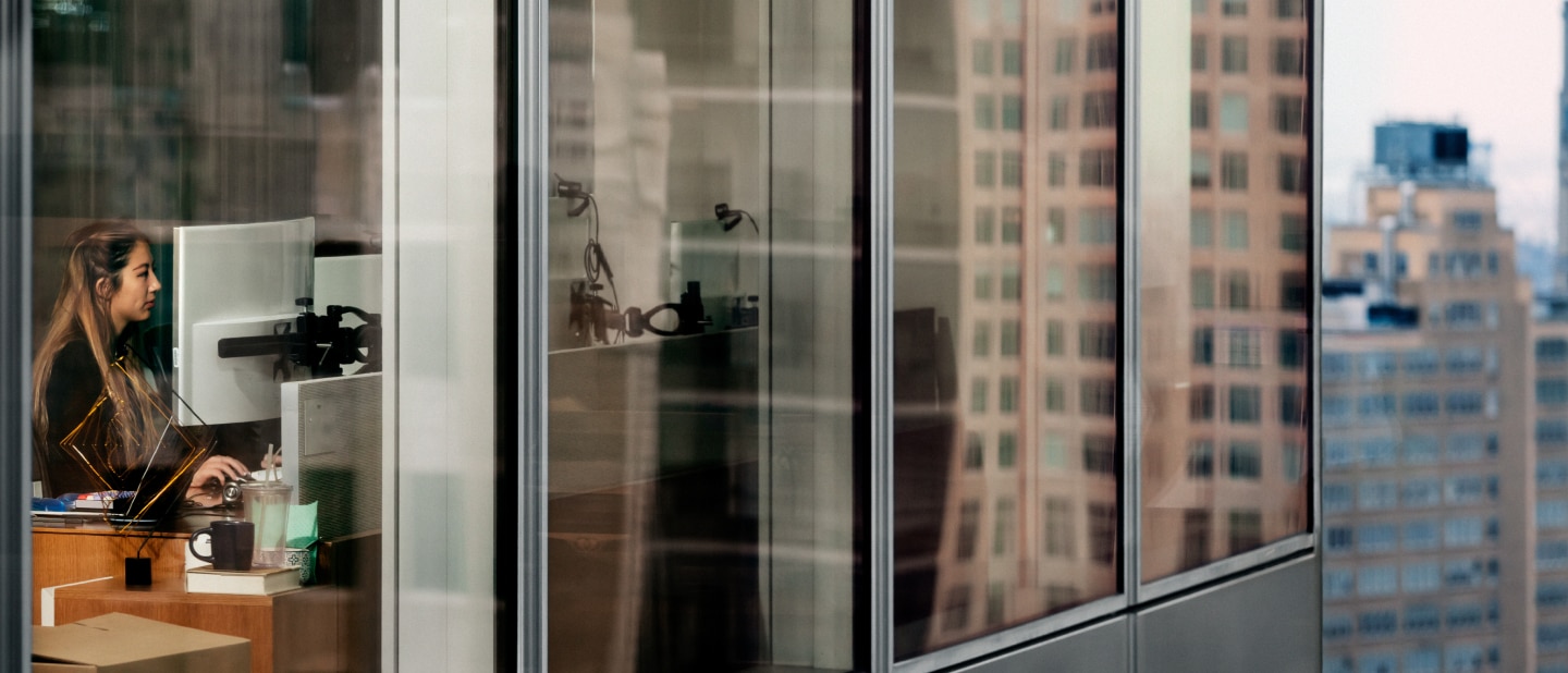 "A woman works at her desk in a modern high-rise office, seen through large glass windows reflecting the surrounding city buildings. The scene captures a blend of interior workspace and urban skyline "