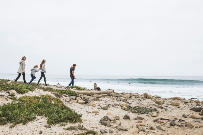 A family walks along a rocky beach path near the ocean, surrounded by sand, greenery, and scattered stones. Waves roll in as surfers are seen in the distance under an overcast sky