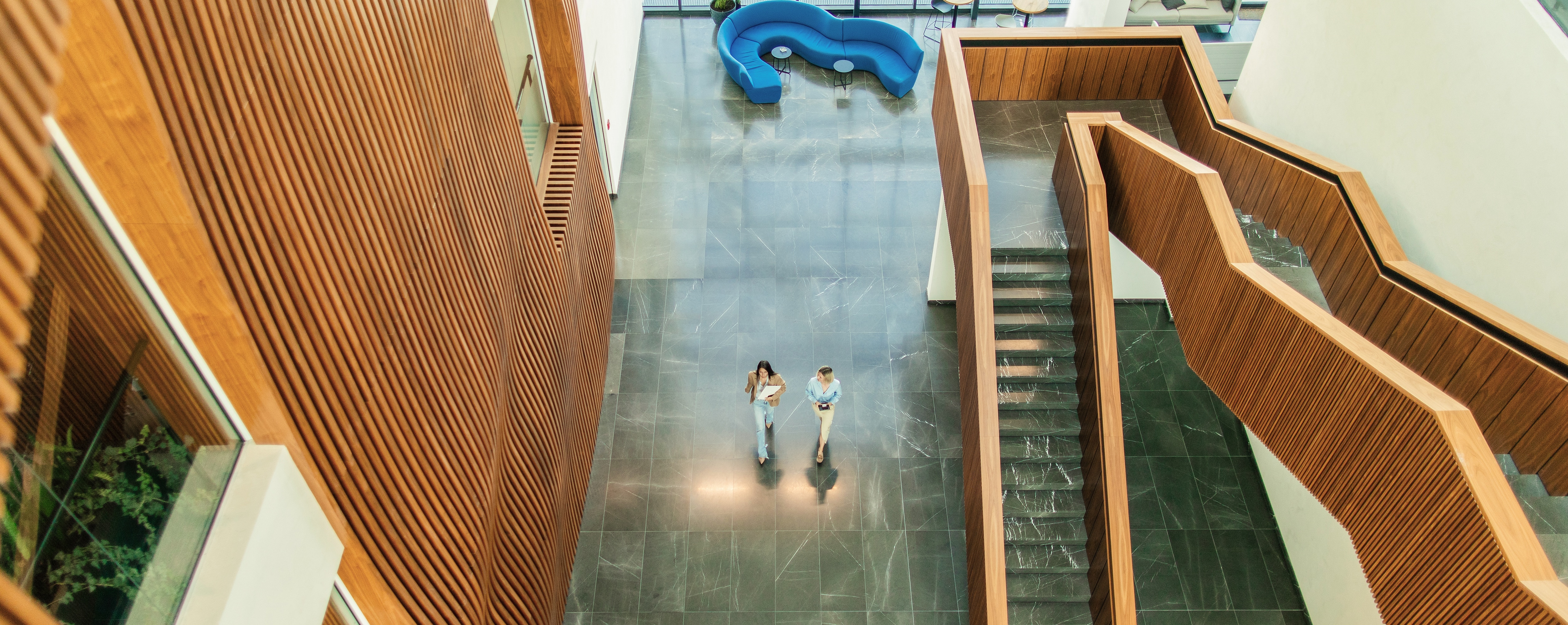 Two people walk through a spacious modern atrium with high ceilings, wooden panel walls, and a geometric staircase. A curved blue seating area is visible in the background
