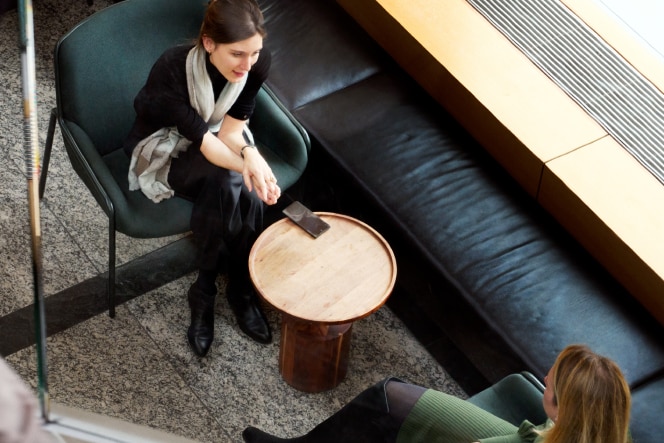 A woman in a black outfit and scarf sits in a modern lounge area, engaged in conversation with another person. The top-down perspective shows a small wooden table, a phone, and surrounding seating in a contemporary setting