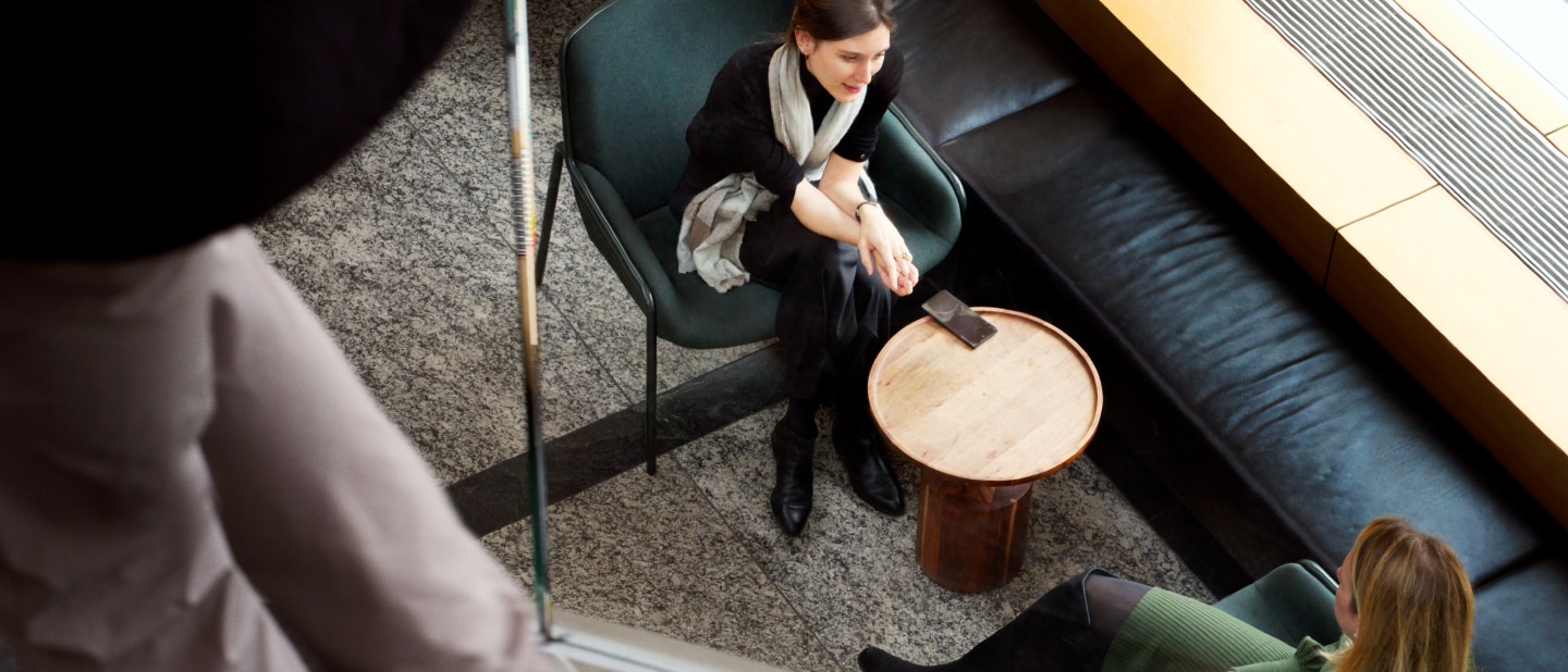 A woman in a black outfit and scarf sits in a modern lounge area, engaged in conversation with another person. The top-down perspective shows a small wooden table, a phone, and surrounding seating in a contemporary setting