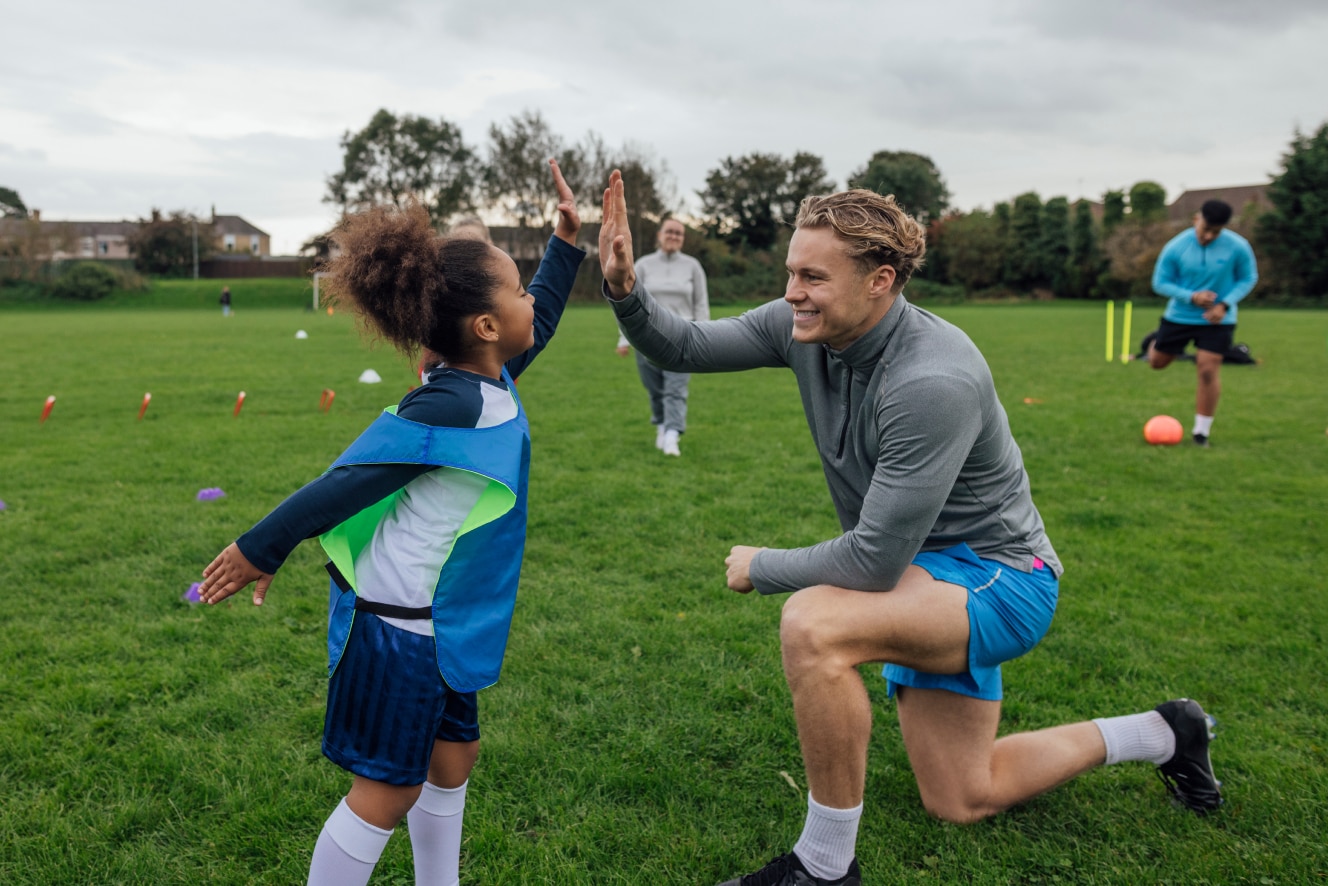 A young girl in a blue sports bib giving a high-five to her smiling coach on a grassy field.