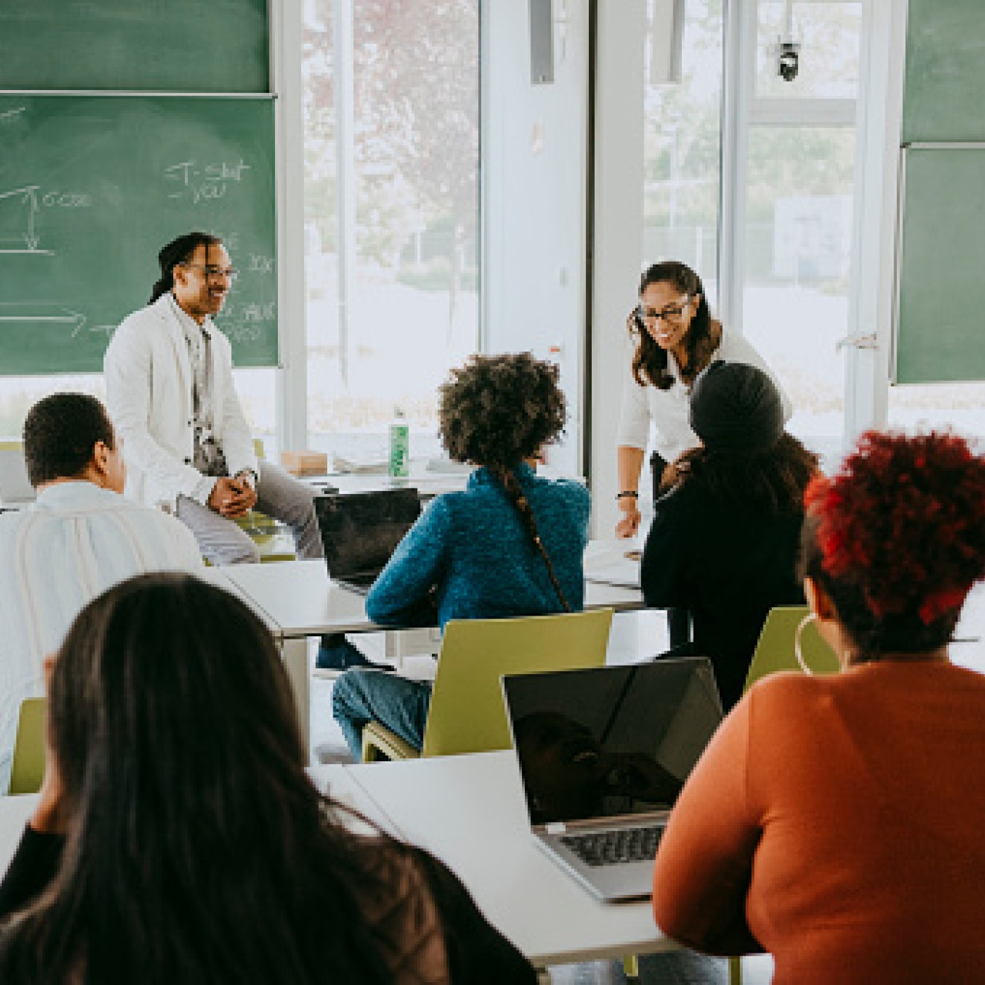 A diverse group of students in a classroom listening to a discussion led by two instructors near a chalkboard.