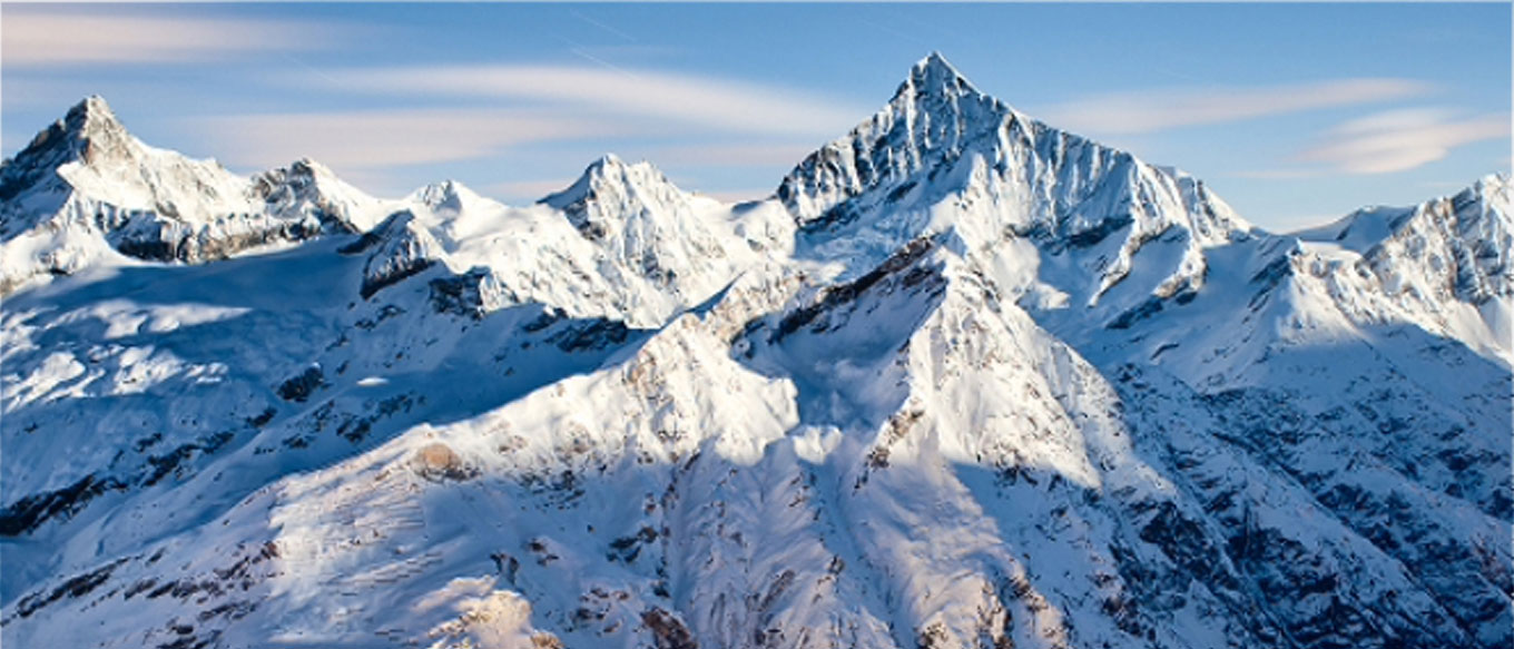 An expansive view of a snowy Alpine mountain range overlooking a valley. The skyline is gradient blue and dotted with wispy white clouds.