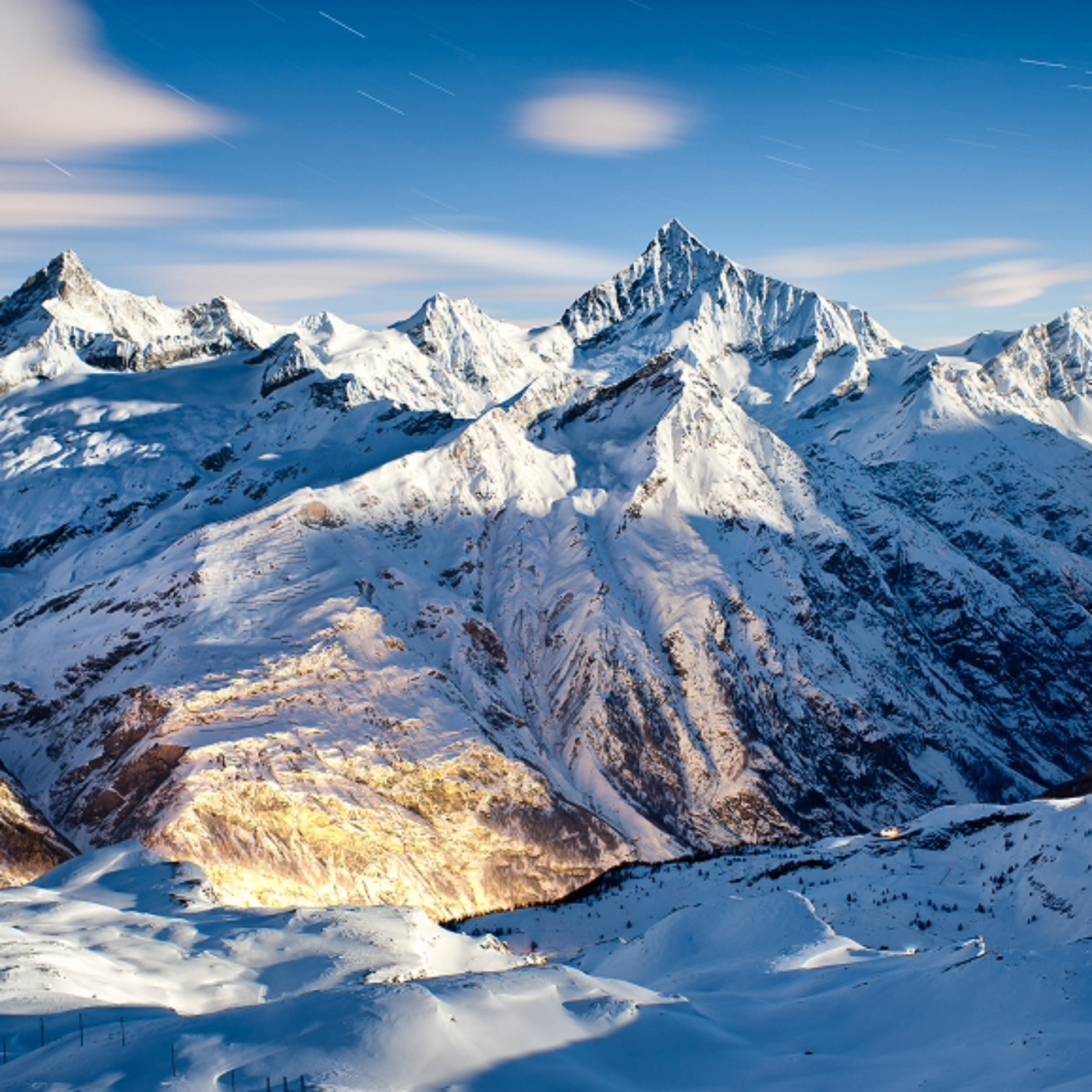 An expansive view of a  snowy Alpine mountain range overlooking a valley. The skyline is gradient blue and dotted with wispy white clouds.