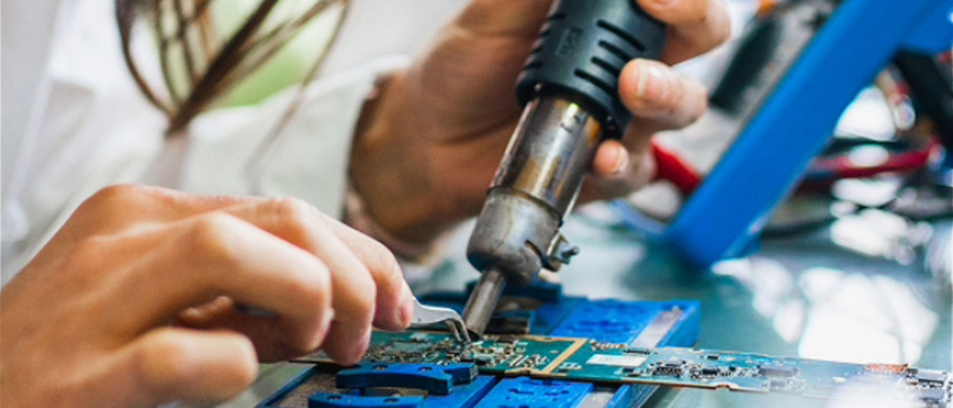 A woman technician meticulously solders components onto a blue and green circuit board with a soldering iron and tweezers. The technician maneuvers the tools with precision.