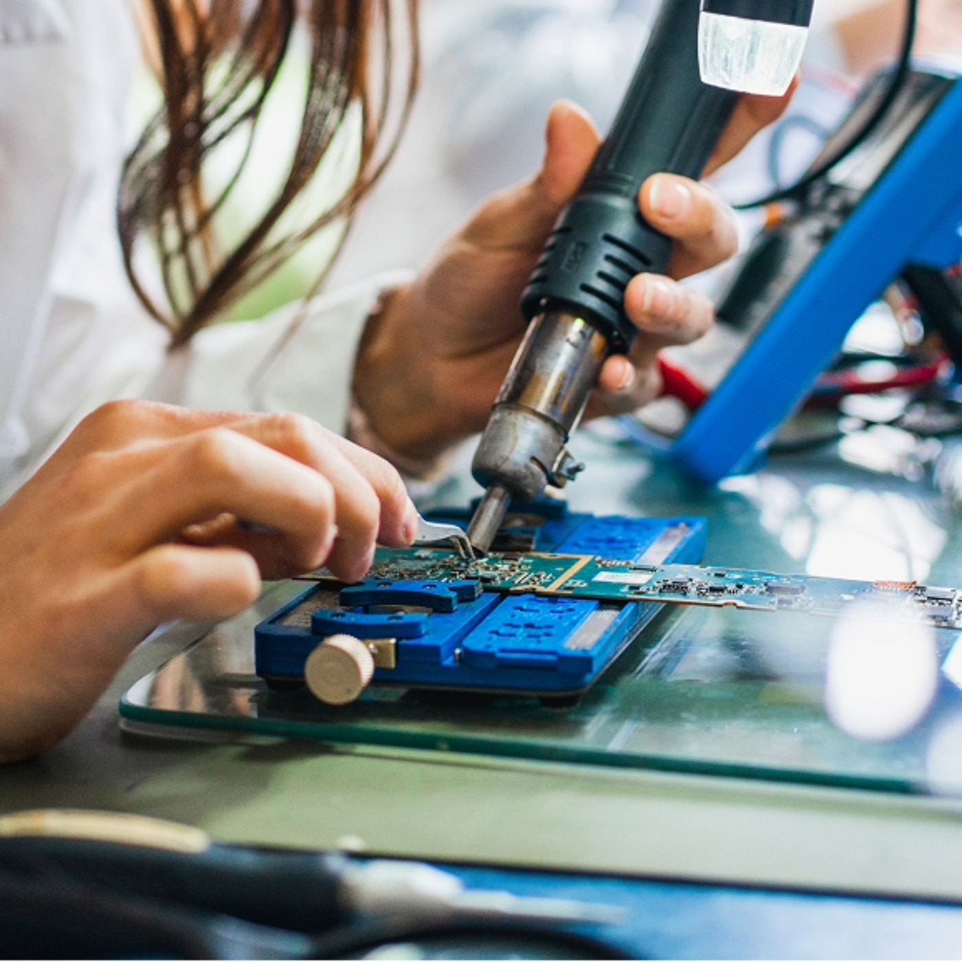 A woman technician meticulously solders components onto a blue and green circuit board with a soldering iron and tweezers. The technician maneuvers the tools with precision.