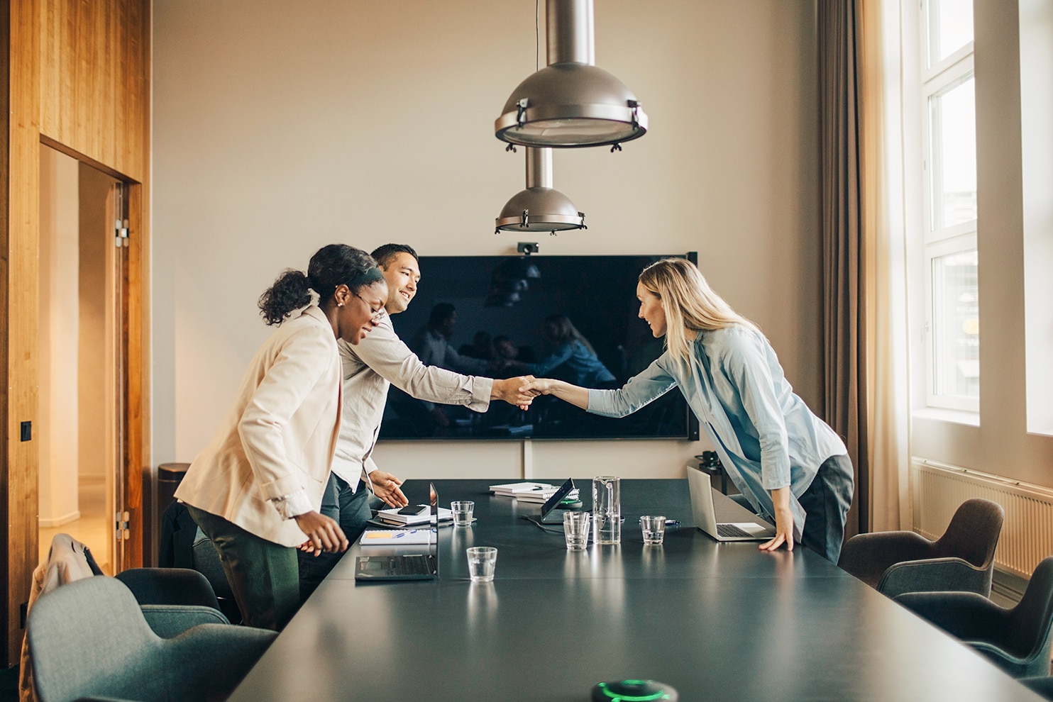 Three colleagues in a conference room smile and shake hands across a table, suggesting a successful meeting or agreement.
