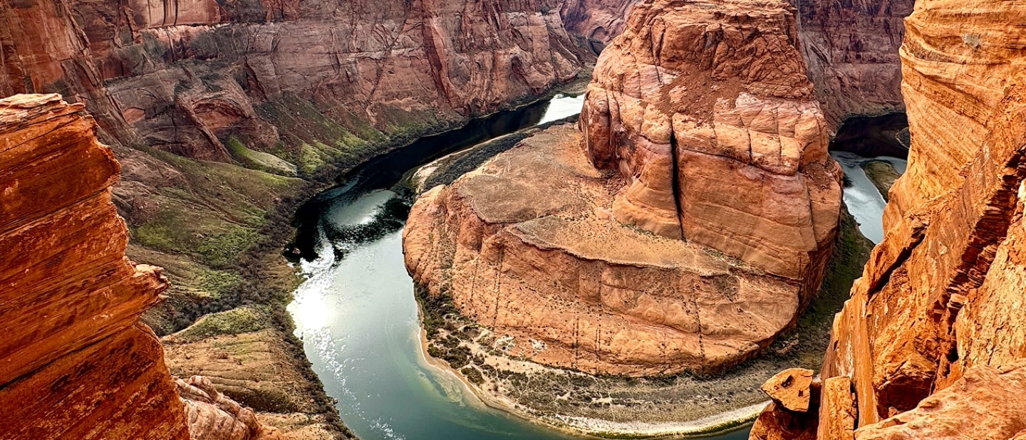 A landscape view of the Grand Canyon’s horseshoe bend on an overcast day.  A shallow river winds around a giant red rock in a horseshoe shape. The rock in the canyon is striped and worn from erosion.
