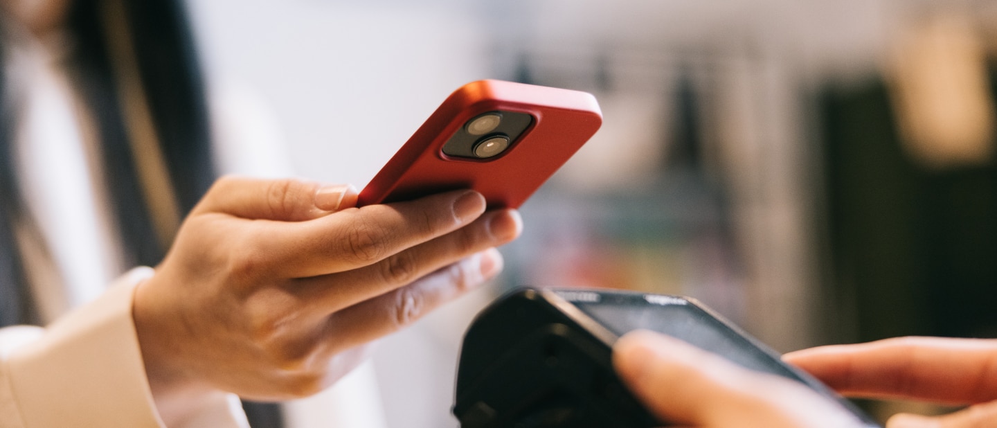 A person holds a red smartphone while making a contactless payment using a card reader. The close-up shot captures the transaction in progress with a blurred background