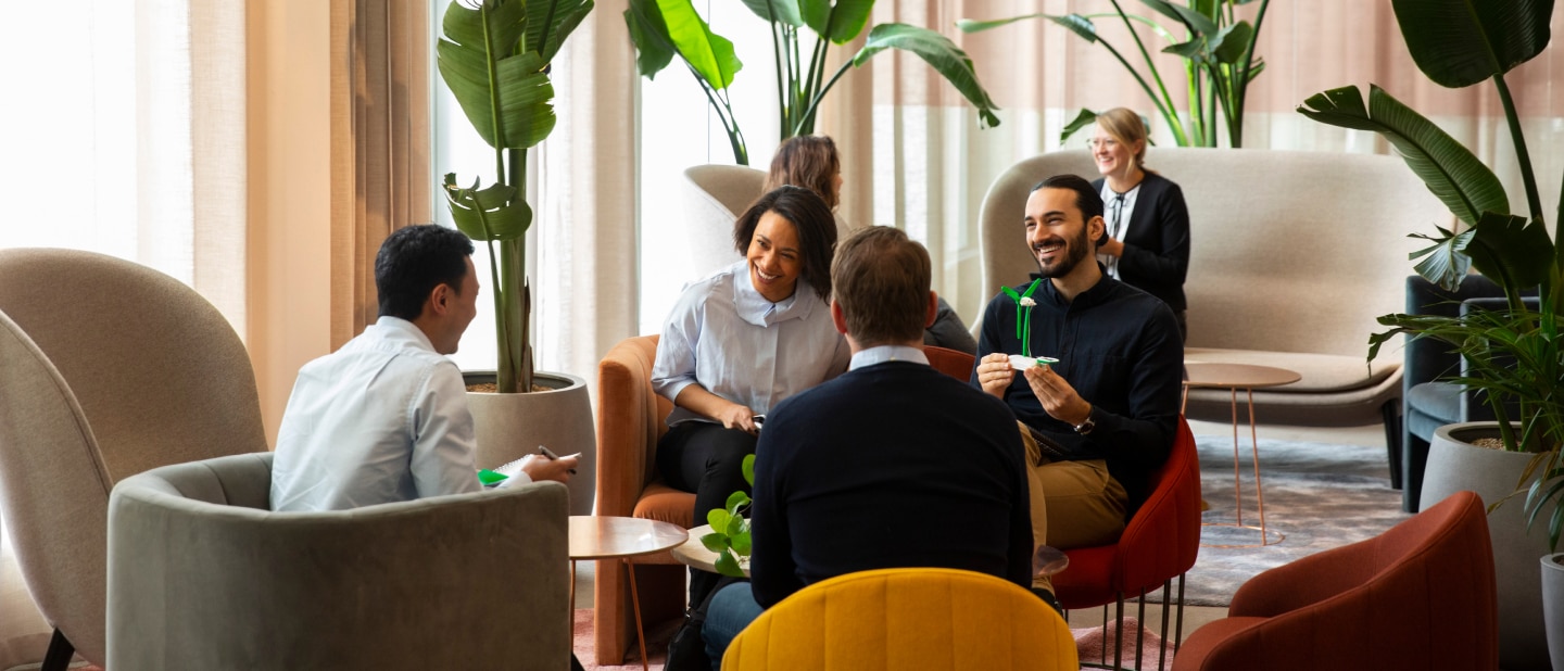 A group of diverse professionals sits in a modern lounge area, engaged in a lively discussion. The space is filled with comfortable seating, greenery, and warm lighting
