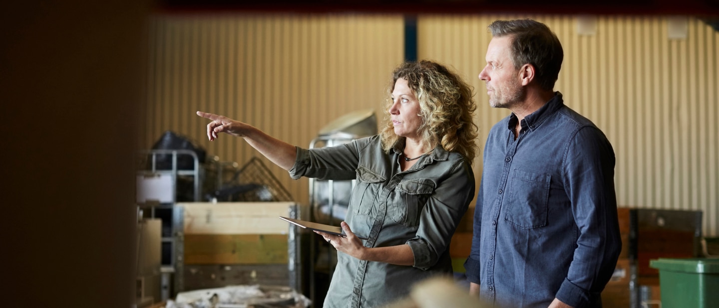 A woman holding a tablet gestures while discussing something with a man in a warehouse-like setting. They both wear casual work attire, surrounded by industrial equipment and storage containers.