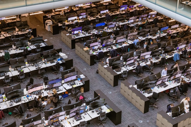 A large open-plan trading floor with numerous desks equipped with multiple computer monitors and office supplies. Employees are seen working at their stations. An elevated glass-walled office section overlooks the trading floor. The scene is illuminated by bright overhead lighting.