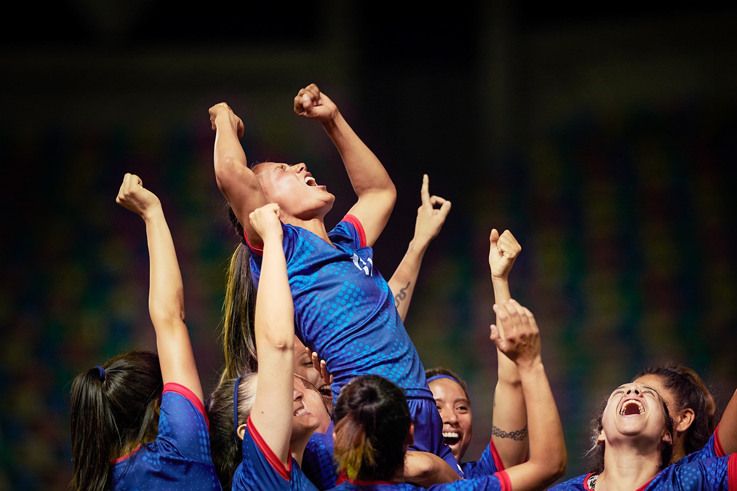 A group of female soccer players in blue and red uniforms celebrating a victory. They lift a teammate into the air, who shouts with joy, surrounded by teammates cheering and raising their arms in triumph.