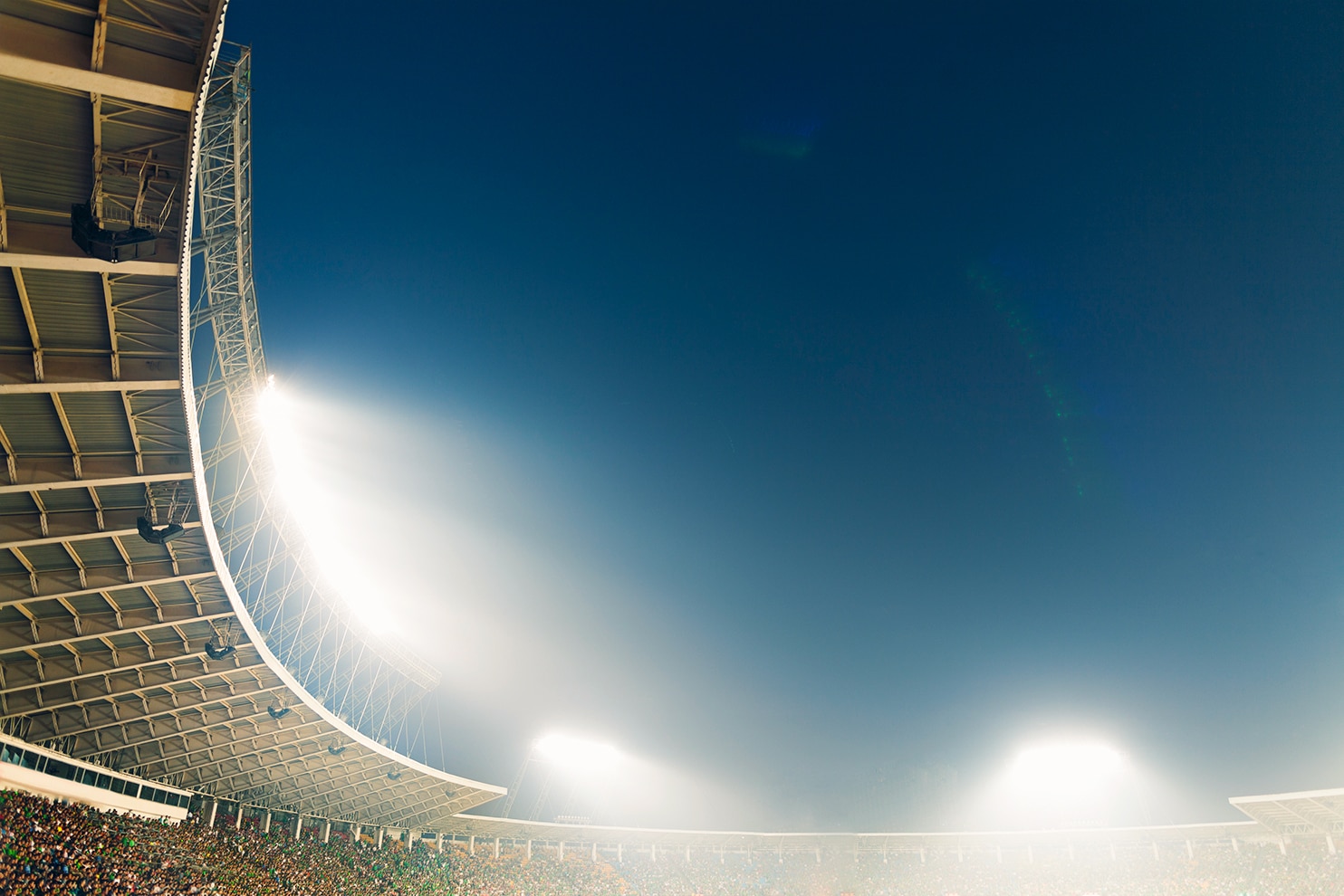 A stadium scene at night, featuring towering floodlights illuminating the crowd and structure. The camera angle captures the vastness of the venue, with the curved roof and bright lights against a deep blue sky.