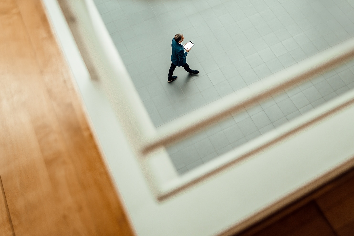 A man in a blue blazer walks across a tiled floor, holding a tablet, viewed from above through a railing. The perspective creates a framed effect, emphasizing movement and space