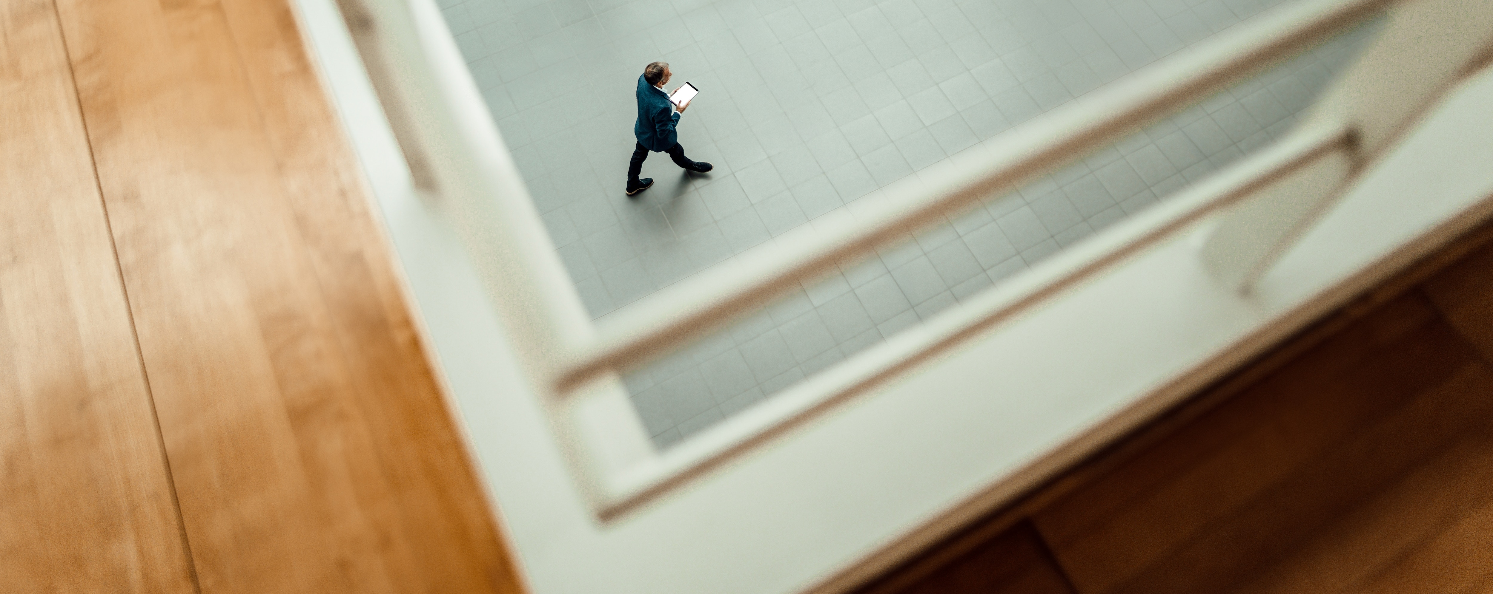 A man in a blue blazer walks across a tiled floor, holding a tablet, viewed from above through a railing. The perspective creates a framed effect, emphasizing movement and space