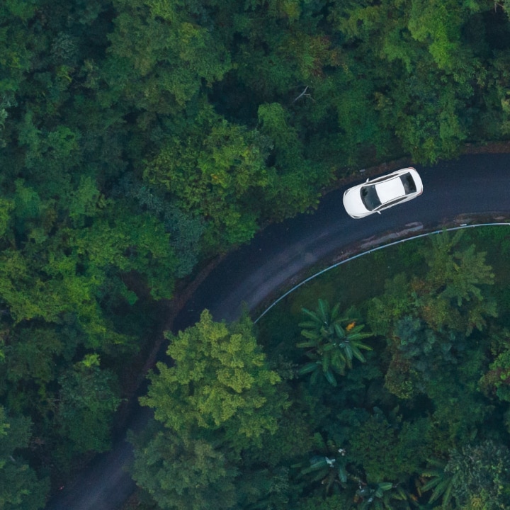 A white car driving on a winding road surrounded by lush green forest, captured from a bird's-eye view.