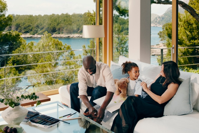 A family relaxes in a bright, modern living room with floor-to-ceiling windows overlooking a scenic coastal view. A man and a young girl are drawing together while a woman watches, smiling. The space is filled with natural light, greenery, and cozy furnishings.
