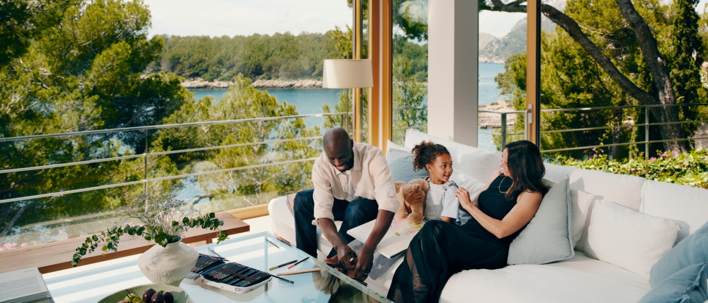 A family relaxes in a bright, modern living room with floor-to-ceiling windows overlooking a scenic coastal view. A man and a young girl are drawing together while a woman watches, smiling. The space is filled with natural light, greenery, and cozy furnishings.