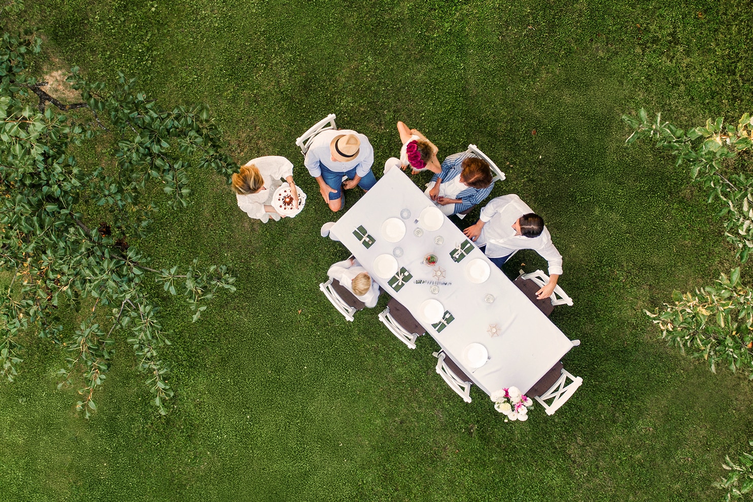 A group of people gathers around a long, white-clothed table set for a meal in a lush green garden. The aerial view captures the setting among trees